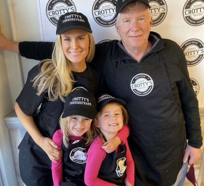 Lindsay Crotty Walsh with her dad Joe Crotty and two daughters in their custom apparel Smiling blonde woman smiling man, and two little girls all wearing Crotty's custom hats, custom t-shirts, and custom aprons stand in front of a wall of Crotty's custom logos.