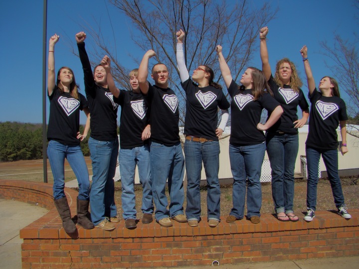 A close squad of friends wears matching custom shirts and raises their hands like superman flying.